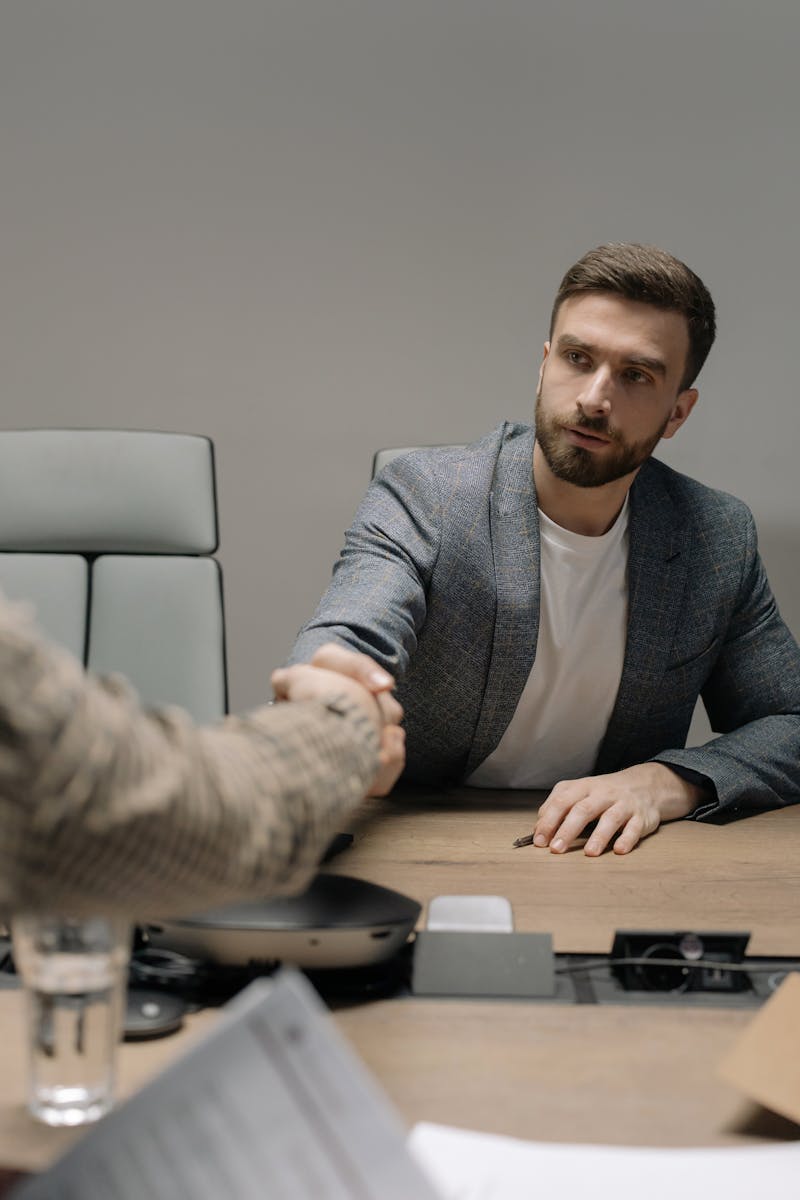 Two businessmen in a modern office shaking hands across a table. Professional setting.